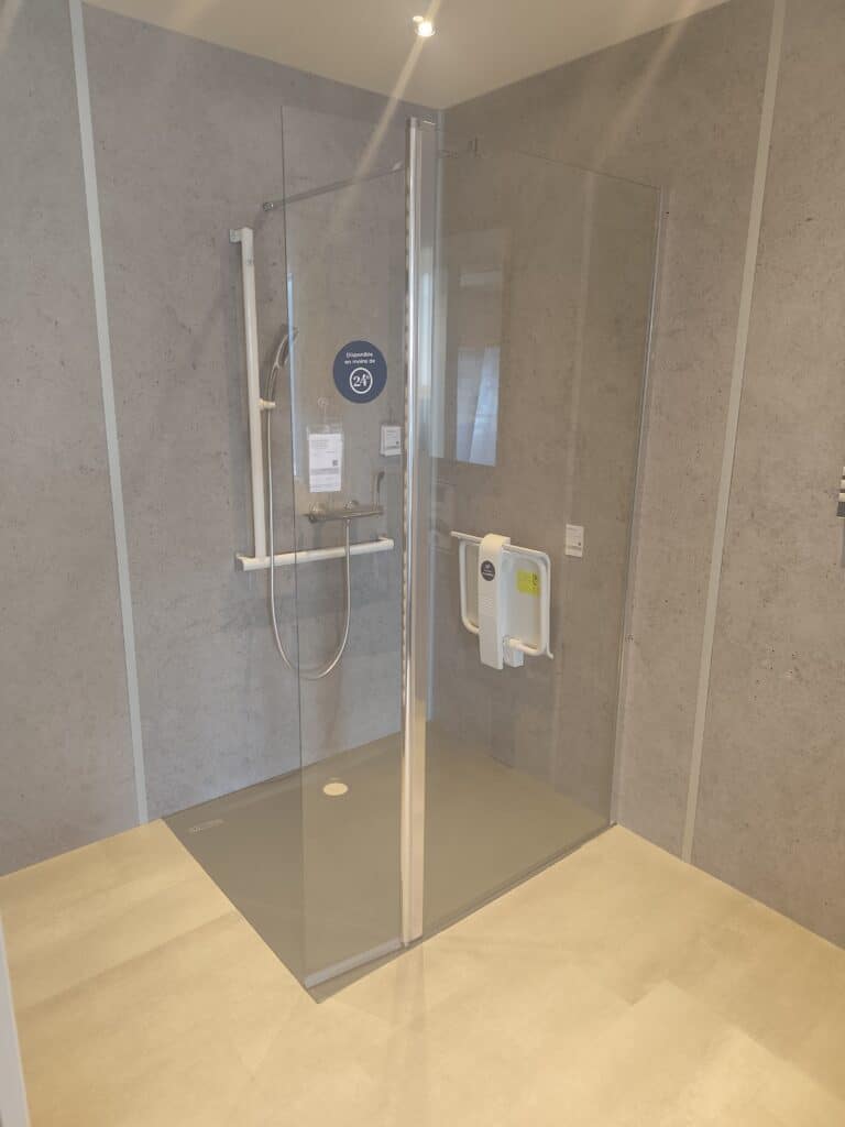Aménagement Douche PMR Sécurisée avec Siège Escamotable Douche PMR avec paroi vitrée, barre de maintien, siège rabattable et receveur gris. Disponible en moins de 24h.
