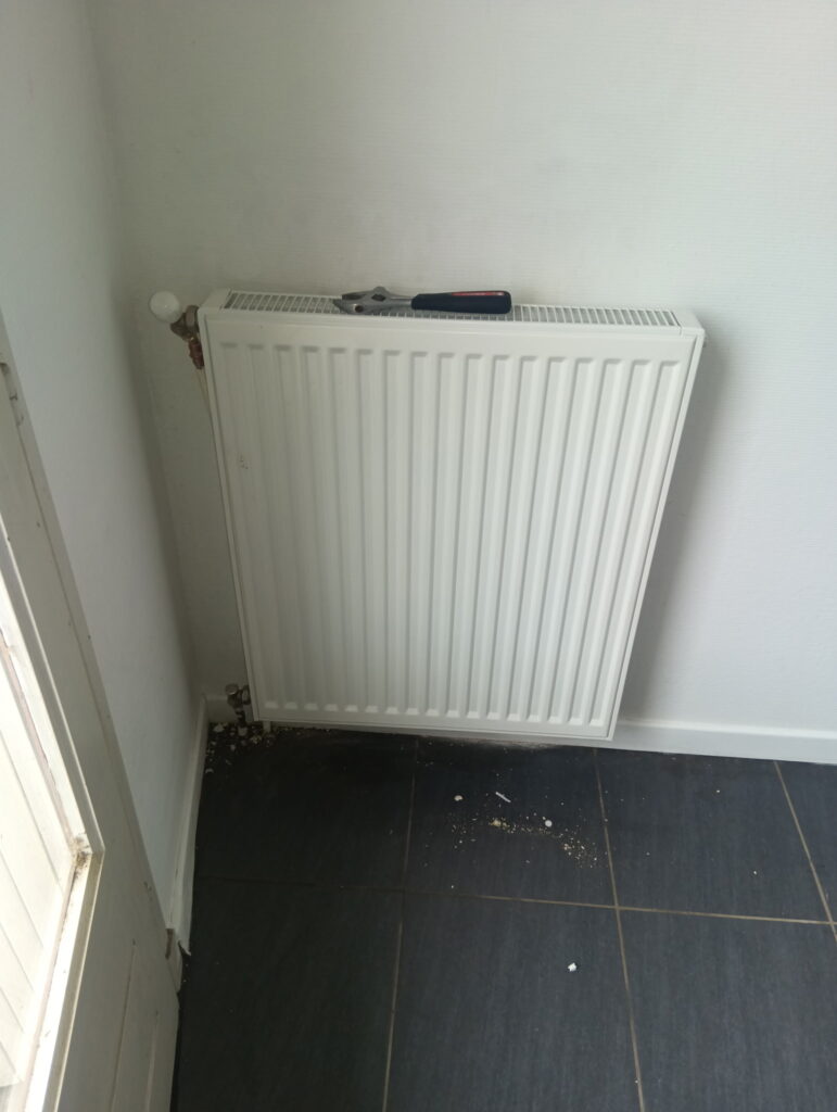 Radiateur blanc mural avec une clé à molette, posé sur un carrelage gris foncé dans le coin d'une pièce.