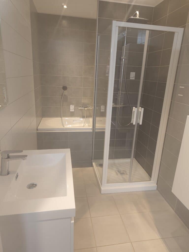 Aménagement Salle de Bain : Douche Italienne et Baignoire Salle de bain moderne avec baignoire, douche à l'italienne vitrée et lavabo blanc sur carrelage gris.